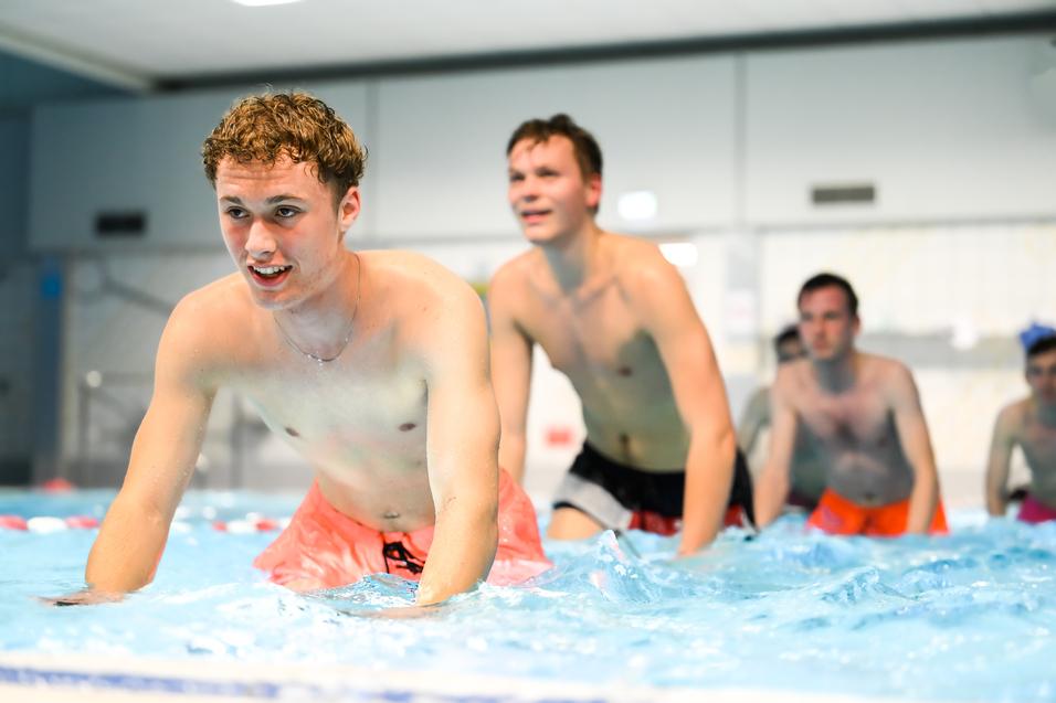 Vier junge Männer in Badebekleidung üben im Wasser eines Schwimmbeckens, einige lachen und spritzen.